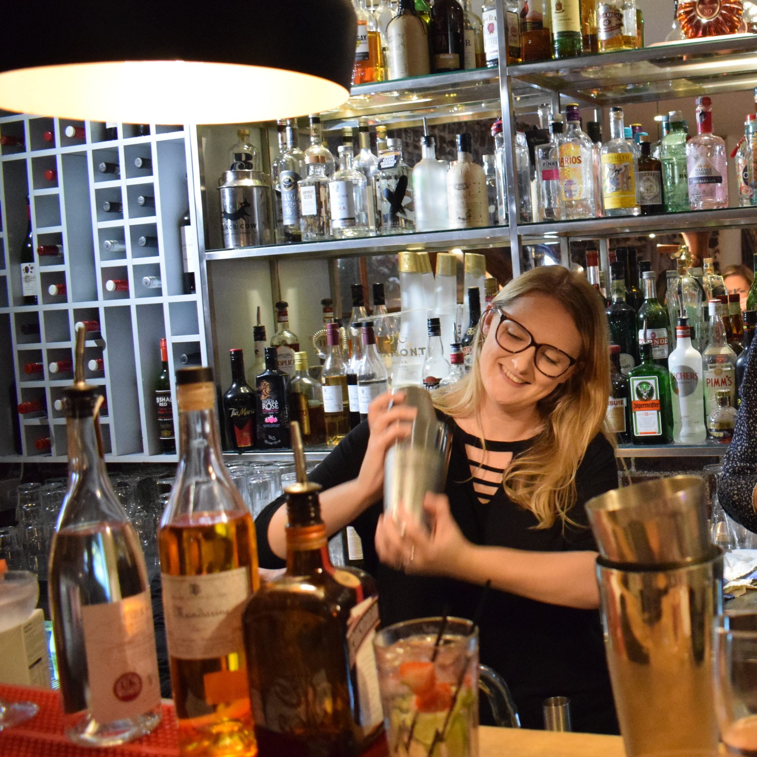 Photo shows a blonde woman in her 20s standing behind a bar, shaking a cocktail maker and smiling.